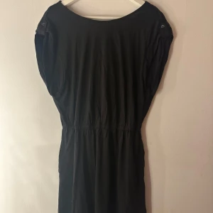 Svart kortklänning med knappar. Levis  - Svart kortklänning i mjuk bomull med resår i midjan och snygga knapprankor på axlarna. Klänningen har lös passform och ärmlös design med lätt draperad rygg. Perfekt för dig som gillar enkel och stilren look. Levis. Storlek M