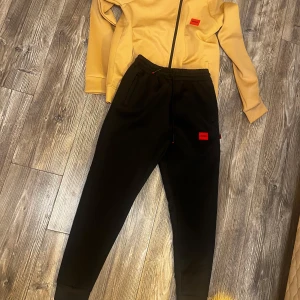 Hugo tracksuit - Snygg beige vindjacka från Hugo med svart dragkedja och huva. Jackan har en röd Hugo-logga på bröstet och är tillverkad i ett mjukt syntetmaterial. Perfekt för dig som gillar stilrena och sportiga plagg med en modern touch. 