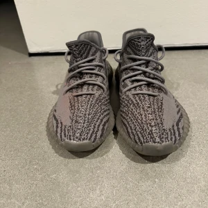 Adidas Yeezy Boost 350 V2 Beluga - Säljer ett par Adidas Yeezy Boost 350 V2 Beluga med grå och mörkgrå stickad ovandel, orange SPLY-350-tryck på sidan och en chunky, genomskinlig sula. Skorna har snörning och en unik mönstrad design som sticker ut.