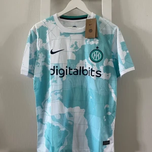 Inter Milan bortatröja 2024 Nike M - Inter Milans officiella bortatröja 2024 från Nike. Tröjan är vit med ljusblått världskartemönster, svart Nike-logga och Inter-emblem på bröstet. Digitalbits och Lenovo som sponsors. Tillverkad i lätt och ventilerande material, perfekt för match eller träning.