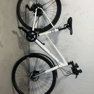 Välvårdad vit hybridcykel – snabb, lätt och snygg! - Bilderna visar en vit cykel med svarta detaljer, sportig design och raka styren. Cykeln har skivbromsar, växlar och reflexer, vilket gör den perfekt för både stadskörning och längre turer. Passar dig som vill ha en smidig och modern cykel för vardag och fritid.