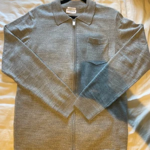 Grå merinoull kofta från Lindbergh - Snygg grå kofta från Lindbergh i mjuk merinoull. Modellen har hel dragkedja, klassisk krage och en bröstficka. Slim fit-passform och långa ärmar gör den perfekt för lager-på-lager. Enkel och stilren look som funkar året runt. Frågor är de bara att kontakta!