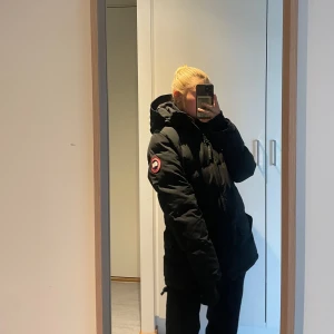 Svart dunjacka från Canada Goose M - Säljer en äkta svart dunjacka från Canada Goose med huva och klassisk logga på ärmen. Jackan har dragkedja framtill, stora fickor och en clean look. Perfekt för kalla dagar och riktigt snygg till streetwear-stil.