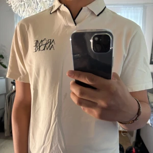 Vit piké från H&M med tryck - Snygg vit pikétröja från H&M med svart text 'ARÔME AMORE' på bröstet. Klassisk krage med svarta detaljer och kort ärm. Regular fit och tillverkad i bomull, perfekt för en clean och stilren look.