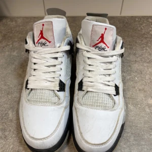 Nike Air Jordan 4 White Cement - Nike Air Jordan 4 White Cement med vit ovandel i skinn, grå och svarta detaljer samt röd Jumpman-logga på plösen. Snygg mesh på sidorna och klassisk Nike Air-logga på hälen. Skorna har snörning och en robust, svart och grå sula med stänkdetaljer.