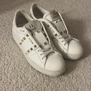 Valentino Rockstud vita sneakers - Snygga vita Valentino Rockstud sneakers i skinn med ikoniska guldiga nitar längs sidorna och bak på sulan. Klassisk rund tå, platt sula och snörning framtill. Perfekta för dig som vill ha en lyxig och edgy touch till din outfit. Använda ca ett halvår! 😇