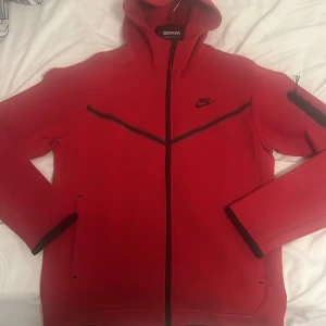 Röd Nike Nike tech - Nike vindjacka i rött med svarta detaljer och dragkedja framtill. Jackan har huva, långa ärmar och två fickor. Perfekt för dig som gillar sportig stil och vill sticka ut med en färgstark look.