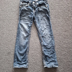 True Religion jeans med kontrastsömmar - Säljer ett par blå jeans från True Religion med tydliga vita kontrastsömmar och stora fickor med lock och knappar bak. Klassisk femficksmodell med silverfärgade knappar och logopatch bak. Raka ben och cool tvättad look.