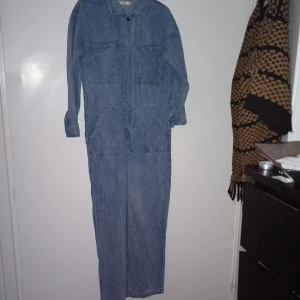 Blå jeans jumpsuit med fickor - Säljer en blå jeans jumpsuit med långa ärmar och stora fickor både fram på bröstet och vid höfterna. Klassisk skjortkrage och stängning framtill. Perfekt för dig som gillar en avslappnad och cool stil.