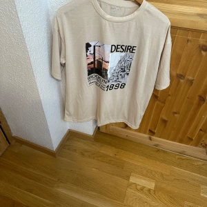 Beige t-shirt med Brooklyn-tryck från SHEIN - Beige t-shirt från SHEIN med coolt tryck framtill som visar Brooklyn och texten 'DESIRE 1898'. T-shirten har rund hals, korta ärmar och en avslappnad passform. Perfekt för dig som gillar streetstyle och snygga grafiska prints.