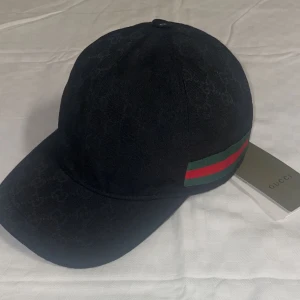 Svart Gucci keps - Svart keps från Gucci med diskret GG-logomönster i tyget och klassiska gröna och röda ränder på sidan. Justerbar rem baktill i skinn. Perfekt för dig som vill ha en stilren och lyxig accessoar.