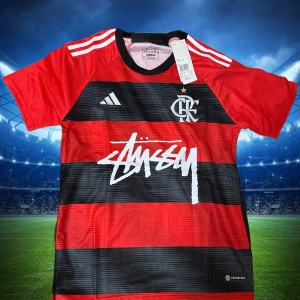 Randig Flamengo x Stüssy Adidas tröja - Snygg fotbollströja från Adidas i samarbete med Flamengo och Stüssy. Tröjan är röd och svart med breda horisontella ränder, korta ärmar och vita detaljer. Tillverkad i lätt och ventilerande material med klubbmärke och Stüssy-logga på bröstet.