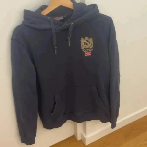 Mörkblå hoodie från Morris, storlek S - Säljer en mörkblå hoodie från Morris med guldfärgat broderat emblem och brittisk flagga på bröstet. Tröjan har huva med snörning och en stor magficka. Perfekt för dig som gillar klassisk stil med en twist.