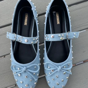 Ljusblå ballerinas med nitar från Cushionaire - Snygga ljusblå ballerinas från Cushionaire i jeansmaterial med silverfärgade nitar och dekorativ rosett framtill. Skorna har en rem över foten med spänne och rund tå. Ej använda men finns lite slitning på sidan av skorna på bild fyra 