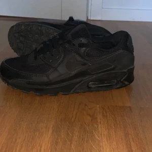 Svarta Nike Air Max  - Säljer ett par helsvarta Nike Air Max sneakers med klassisk siluett. Skorna har mesh och skinnpaneler, synlig Air-dämpning i sulan och snörning framtill. Perfekta för dig som gillar en clean och sportig look.