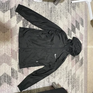 Nike vindjacka med storlek Small - Svart vindjacka från Nike med klassisk vit swoosh på bröstet. Jackan har huva, dragkedja framtill och två fickor med dragkedja. Tillverkad i lätt polyester som passar perfekt när det blåser. Enkel och stilren design som funkar till allt.