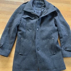 Mörkgrå rock från Selected Homme -  Obs!!! Om ni köper så köp via PostNord annars kan jag inte skicka denna rock! Säljer denna rock för detta pris eftersom att som ny kostar den 2750 kr. Det är en stilren mörkgrå rock från Selected Homme med klassisk krage, knappar framtill och två fickor med knappdetaljer. Jackan har lång ärm och är tillverkad i ullblandning, perfekt för kyliga dagar. Snygg passform och tidlös design som funkar till många outfits. Den är i storlek M.