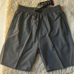 Grå träningsshorts från Under Armour - Snygga grå träningsshorts från Under Armour med elastisk midja och dragsko. Shortsen har diskreta loggor på benet och bak, samt praktiska sidofickor. Perfekta för gymmet eller löprundan, tillverkade i lätt och snabbtorkande syntetmaterial.