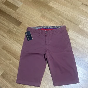 Vinröda shorts från Pitbull, stl 30 - Snygga vinröda shorts från Pitbull i bomull med lite stretch. Klassisk modell med bälteshällor, fickor fram och bak samt dragkedja. Insidan har ett coolt mönstrat tyg som ger extra stilpoäng. Perfekta för sommaren när du vill vara både bekväm och trendig.