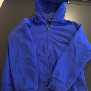 Blå hoodie från Polo Ralph Lauren - Blå hoodie från Polo Ralph Lauren med dragkedja och huva. Klassisk röd broderad logga på bröstet. Mjuk och skön i bomullsmix, perfekt för chill dagar. Långärmad och med ribbade muddar för extra komfort. Kan göra byten. 14-16 använd nån gång 