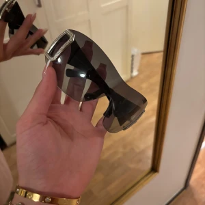  solglasögon med strassdetaljer - Snygga svarta solglasögon från D&G med stora, mörka glas och breda skalmar. På sidorna finns en stor G-logga i silver med glittrande strass. Glasögonen har en futuristisk vibe och passar perfekt om du vill sticka ut.