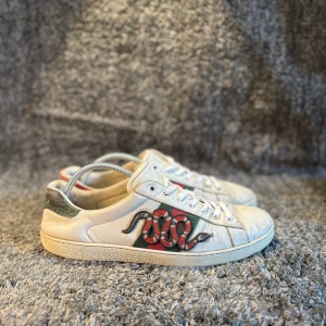 Gucci skor  - Snygga vita sneakers från Gucci i skinn med broderad röd och svart orm på sidan, gröna och röda detaljer samt häftig ormskinnslook på hälen. Klassisk rund tå, snörning och platt sula. Perfekta för dig som vill sticka ut med lyxig streetstyle.