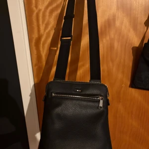 Hugo boss väska - Säljer en stilren svart axelväska i skinn med justerbar rem och praktisk dragkedja framtill. Perfekt storlek för att få plats med det viktigaste och passar till många olika outfits. Enkel och clean design som funkar till det mesta.