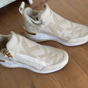 Beige sneakers från Michael Kors med gulddetaljer - Säljer ett par snygga beige sneakers från Michael Kors med stora MK-loggor i tyg och gulddetaljer vid dragkedjan och hälen. Skorna har vit och brun sula samt dragkedja på sidan för enkel påtagning.