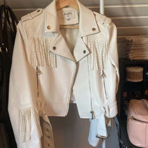 Vit skinnjacka med fransar Soy&Co - Snygg vit skinnjacka från Soy&Co med coola fransdetaljer och nitar på bröstet och ärmarna. Jackan har dragkedjor, bälte i midjan och axelklaffar. Perfekt för dig som vill sticka ut med en edgy look.