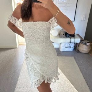Vit spetsklänning med volangdetaljer - Säljer en söt vit kortklänning med spets och volangdetaljer. Klänningen har offshoulder-ärmar och en omlottliknande kjol med volangkant. Perfekt för sommarens tillfällen! Aldrig använd med prislapp på!