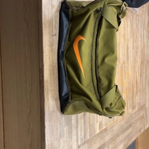 Olivgrön träningsväska från Nike - Säljer en olivgrön träningsväska från Nike med orange logga på sidan och axelrem. Väskan har dragkedja och flera fack, perfekt för gymmet eller sportaktiviteter. Snygg och praktisk design med plats för allt du behöver.