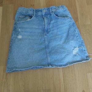 Ljusblå jeanskjol med slitningar - Säljer en ljusblå jeanskjol med slitna detaljer framtill och klassiska fickor både fram och bak. Kjolen har en rak passform och stängs med knapp och dragkedja. Perfekt för en avslappnad stil.