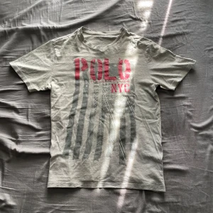 Grå t-shirt från Polo Ralph Lauren - Säljer en unik grå t-shirt från Polo Ralph Lauren med stort rött 'POLO NYC'-tryck och grafiskt motiv framtill. Perfekt för en avslappnad stil. Pris kan diskuteras