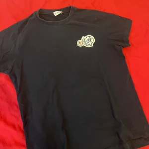 Mörkblå t-shirt från Moncler med logga - Säljer en mörkblå t-shirt från Moncler med klassisk rund hals och Moncler-logga på bröstet. Enkel och stilren design som passar perfekt till jeans eller shorts. T-shirten är i mjuk bomull och har korta ärmar.
