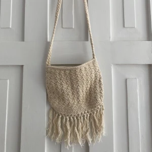 Beige axelväska med fransar från Indiska - Säljer en beige axelväska från Indiska i virkat material med långa fransar nedtill. Väskan har en flätad axelrem!