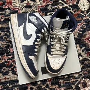 Nike Air Jordan 1 Mid Navy/Vit - Säljer ett par Nike Air Jordan 1 Mid i marinblått och vitt. Skorna har klassisk hög siluett, vit bas med mörkblå overlays och vit sula. Ikoniska Air Jordan-loggan på sidan och Jumpman på plösen. Perfekta för dig som gillar streetwear och sneakers.