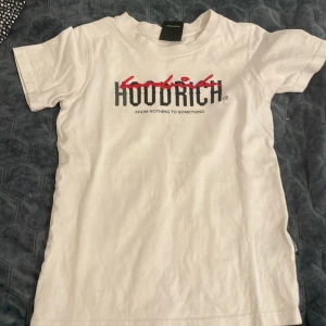 Två Hoodrich t-shirts i olika färger - Säljer två snygga t-shirts från Hoodrich. En är helvit med tryck framtill och en är svart och grå med röda detaljer och stor logga på bröstet. Båda har rund hals och korta ärmar, perfekta för en streetwear-look.