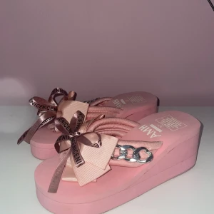 Rosa platåsandaler med rosett och kedjedetalj - Säljer ett par rosa platåsandaler från AMH med stora rosetter och textband, samt en silvrig kedjedetalj på remmen. Skorna har öppen tå och hög platåsula, perfekta för dig som gillar statement-detaljer. aldrig använt 