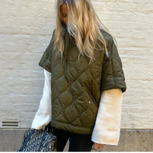 Olivgrön quiltad jacka med kort ärm - Säljer en olivgrön quiltad jacka med korta ärmar och halv dragkedja framtill. Jackan har en oversized passform och är tillverkad i syntetmaterial. Perfekt för lager-på-lager och trendig look.