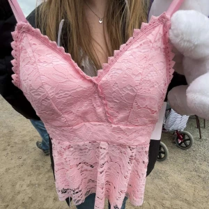 Rosa spetslinne med volangkant - Säljer ett sött rosa linne med spets och volangkant. Linnet har smala axelband och v-ringning, samt ett fint spetsmönster över hela plagget. Perfekt för dig som gillar romantisk stil!