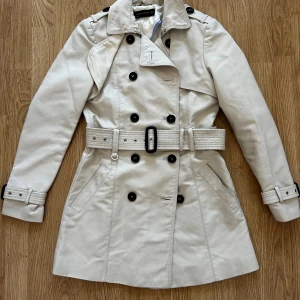 Beige trenchcoat från Zara Woman - Säljer en klassisk beige trenchcoat från Zara Woman med dubbelknäppning, bälte i midjan och svarta knappar. Jackan har långa ärmar, axelklaffar och snygga detaljer vid ärmslut och axlar. Perfekt för en stilren look. Väldigt fint skick dock har sömmen gått upp på insidan.