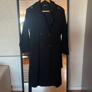 Svart trenchcoat från Gina Tricot - Säljer en svart trenchcoat från Gina Tricot i klassisk modell med knappar och bälte i midjan. Jackan har långa ärmar, axelklaffar och en rak passform. Perfekt för dig som gillar stilrena och tidlösa plagg.