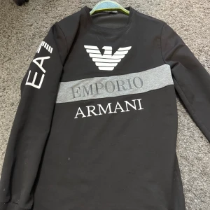 Svart långärmad tröja från Emporio Armani EA7 - Säljer en svart långärmad tröja från Emporio Armani EA7. Tröjan har tryckt logga och text på bröstet samt en grå detalj över bröstet. EA7-tryck på ena ärmen. Perfekt för en stilren och sportig look.