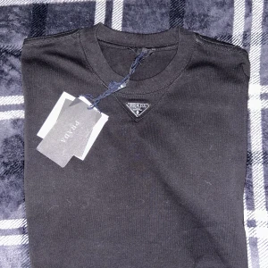 Svart t-shirt från Prada - Säljer en svart t-shirt från Prada med klassisk rund halsringning och den ikoniska metalltriangeln på bröstet. T-shirten är i mjuk bomull och har en stilren, minimalistisk design. Perfekt för dig som gillar exklusiva märken och enkel stil.