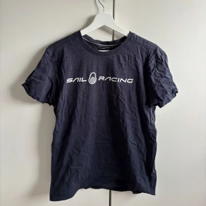 Blå t-shirt från Sail Racing - Säljer en svart t-shirt från Sail Racing med vit logga tryckt på bröstet. Klassisk rund hals och korta ärmar. Perfekt för en avslappnad stil och enkel att matcha med jeans eller shorts.