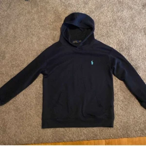 Mörkblå hoodie från Ralph Lauren - Säljer en mörkblå hoodie från Ralph Lauren med klassisk huva och magficka. Tröjan har en liten broderad logga i ljusblått på bröstet och är perfekt för en avslappnad stil.
