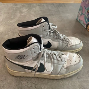 Nike Air Jordans vita och svarta - Säljer ett par klassiska Nike Air Jordans i vitt och svart med högt skaft och svart swoosh. Skorna har snörning och rund tå, samt perforerade detaljer på tån för extra ventilation. Perfekta för dig som gillar streetwear och sneakers.