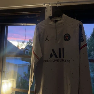 Vit långärmad PSG fotbollströja från Jordan - Säljer en vit långärmad PSG fotbollströja från Jordan med svarta och röda detaljer. Tröjan har klubbmärke på bröstet, Jordan-logga och sponsortryck. Tunn och sportig modell med halv dragkedja och tryck på ärmarna.