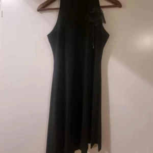 Svart långklänning med blomdetalj och slits - Elegant svart kortlänning med hög hals, dekorerad med små pärlor och en stor tygblomma på axeln. Klänningen är ärmlös och har en slits med pärldetaljer längs kanten. Perfekt för festliga tillfällen. Kan användas som överdel och ha på byxa eller kjol nedan.