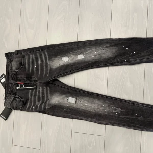 Svarta slitna jeans från Dsquared2 - Säljer ett par svarta jeans från Dsquared2 med slitningar och färgstänk för en cool, edgy look. Byxorna har dragkedja och klassisk femficksdesign. Märket syns tydligt på bakfickan och insidan av linningen. Byxorna är helt oanvända, i bra skick och har alla tags osv
